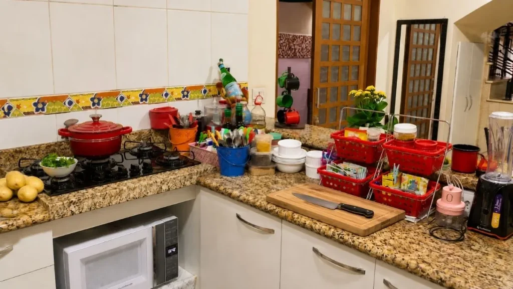 Bancada de cozinha ocupada por utensílios, cestos, potes, eletrodomésticos, tábua de madeira e faca, reduzindo a área livre para preparo dos alimentos.