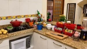 Bancada de cozinha ocupada por utensílios, cestos, potes, eletrodomésticos, tábua de madeira e faca, reduzindo a área livre para preparo dos alimentos.
